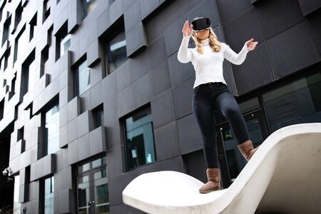 Blonde Woman Wearing Virtual Reality Glasses Against Futuristic Buildingの写真素材
