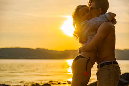 Side view of young man kissing girlfriend standing at beach during summer weekend at beautiful sunsetの写真素材