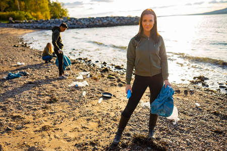 Smiling young woman cleaning beach with volunteers during sunsetの写真素材