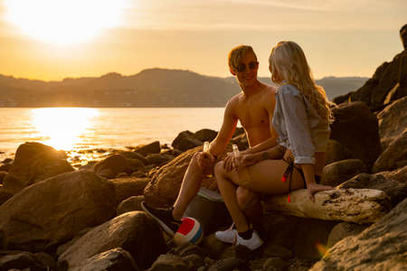 Happy Young Couple Sitting On Rocks Enjoying Beer At Beachの写真素材
