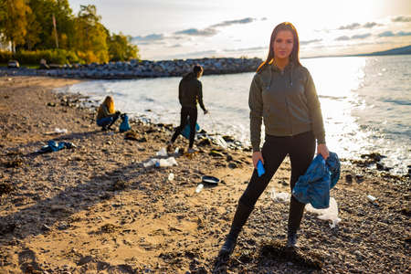 Serious young woman cleaning beach for plastic with volunteers during sunsetの写真素材