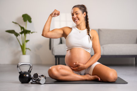 Smiling Strong Pregnant Expecting Mother Showing Off Muscles After Fitness Routine in Living Roomの写真素材