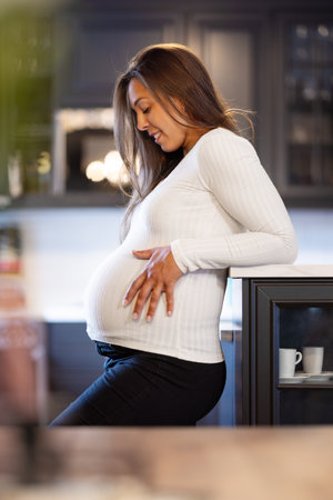 Expectant Smiling Mother Touching Her Pregnant Belly in the Kitchenの写真素材