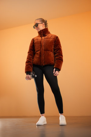 Active woman holding water bottle, wearing trendy jacket and sunglasses indoorsの写真素材