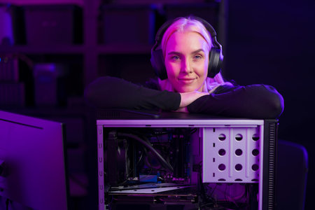 Female gamer with headphones leans on an open gaming PC case under purple lighting, ready to upgrade hardwareの写真素材