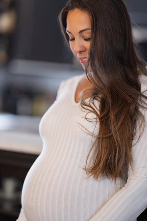 Pregnant Woman in White Attire Contemplating in Serene Home Environmentの写真素材