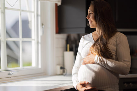 Smiling pregnant woman standing by a window in bright sunlightの写真素材