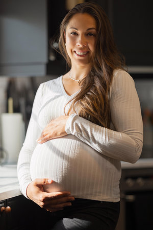 Smiling pregnant woman holding belly in a bright home kitchenの写真素材