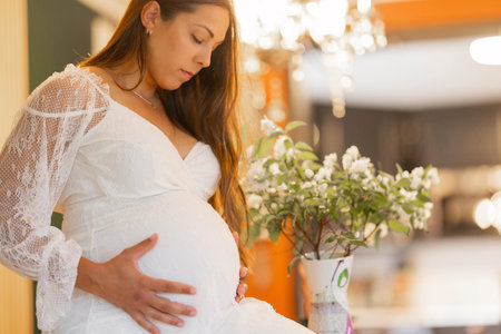 Pregnant woman in white dress holding belly with thoughtful expression indoorsの写真素材