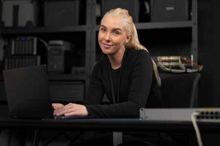 Confident female IT technician at a desk with a laptop and network gear in a technical office