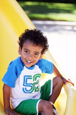A happy little boy with dark, curly hair, playing at a park, on a yellow slide.の写真素材