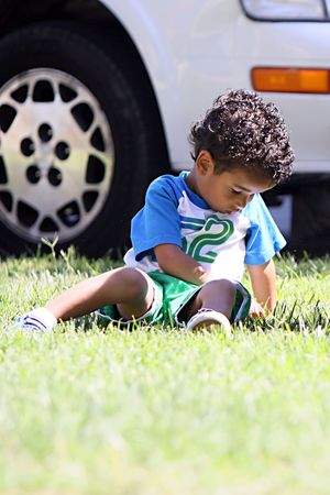 A little boy sitting on the grass picking at it, looking sad.の写真素材