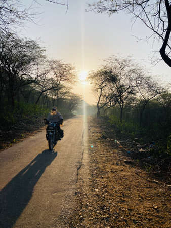 Rear view of a man riding a motorbike on a dirt road at sunsetの写真素材