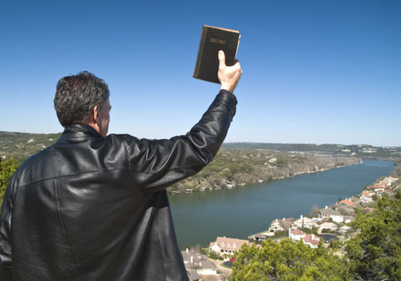 A man holding a bible in his raised hand, looking down over an affluent neighborhood. 