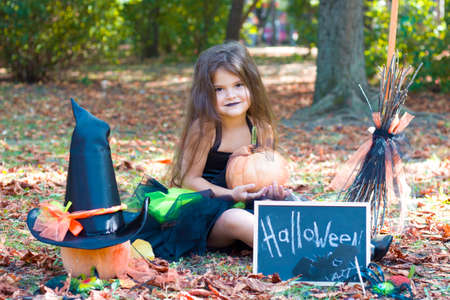 a little girl is sitting in a Park with yellow leaves in a witch costume. Broom, pumpkin, bat mask, witch hat. Halloween.の写真素材