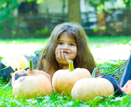 a little girl with black hair in a witch costume is lying on a lawn with pumpkins. Halloweenの写真素材