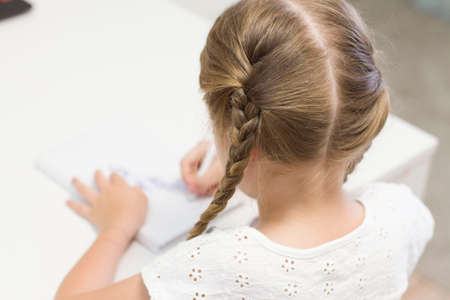 A LITTLE GIRL IN A MASK SITS AT A TABLE AND DRAWS, WRITES. STUDENT. SCHOOL. THE FIRST OF SEPTEMBER.の写真素材