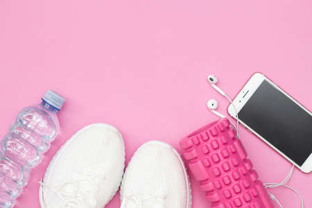 A fitness concept with a water bottle, sneakers, a phone with headphones, a purple fitness elastic band, a pink roller on a pink background. Copy space.の写真素材