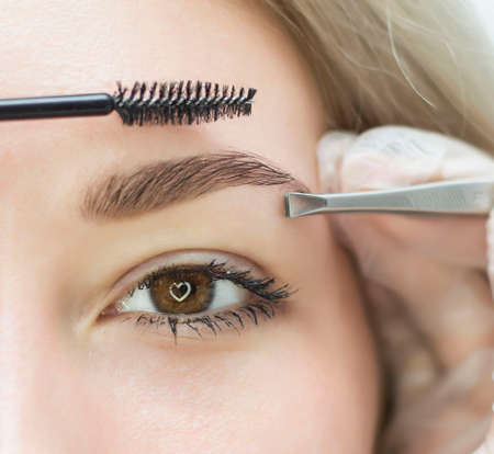 The stylist's hands in white gloves pluck his eyebrows with tweezers. A beautiful attractive female face of a fair-haired well-groomed woman or lady. Styling and lamination of eyebrows.の写真素材
