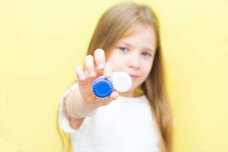 A beautiful girl with long hair holds a container with lenses in her hand. The concept of vision problems in children. Yellow background.の写真素材