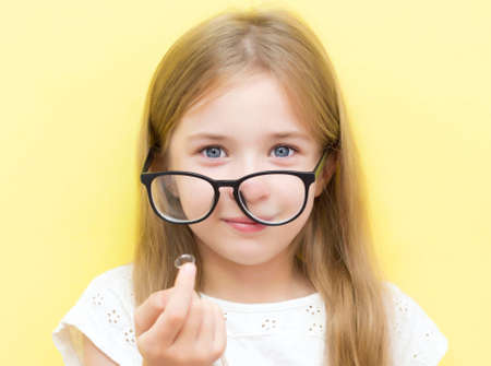A cheerful girl on a yellow background wearing glasses and holding a contact lens. The concept of vision problems in school children.の写真素材
