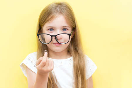 A cheerful girl on a yellow background wearing glasses and holding a contact lens. The concept of vision problems in school children.の写真素材