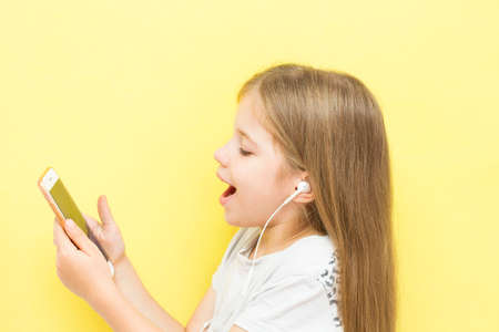 A funny little girl with long hair on a yellow background holds a smartphone in her hands with headphones and smiles. The concept of children and gadgets, social networks, tik tok.の写真素材