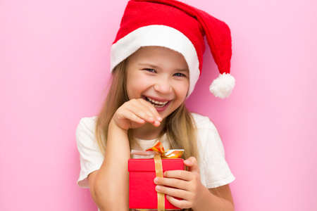 Christmas Eve! A cheerful child in a Christmas hat on a pink background with a red gift box.の写真素材