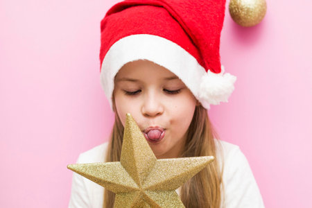 Christmas Eve! A cheerful child in a Christmas hat on a pink background with a gold star in his...