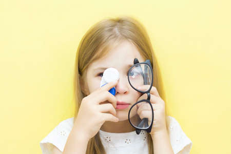 A beautiful baby with long hair holds a container with lenses and glasses in her hands. Choose between glasses and lenses. Yellow background.の写真素材