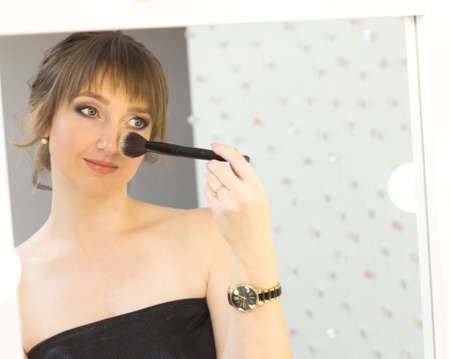 A pretty young lady using a cosmetic brush to apply decorative makeup on her face and looks in the mirror.の写真素材