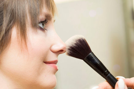 A pretty young lady using a cosmetic brush to apply decorative makeup on her face and looks in the...