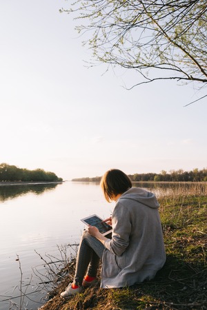 Young girl works with the tablet on the banks of the riverの写真素材