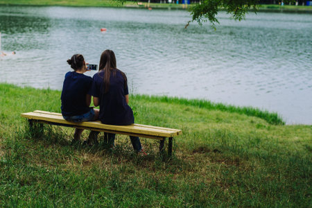 ARMAVIR, RUSSIA - JULE 02, 2016: Young women sitting on a bench by the lake and take pictures of themselvesのeditorial素材