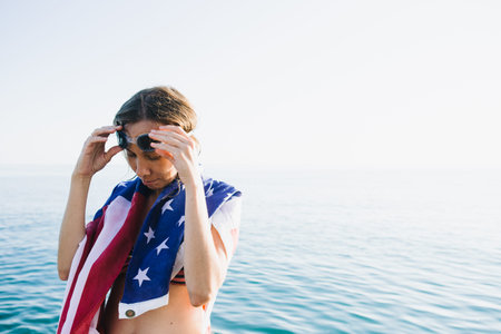 Portrait of young woman in goggles wearing american flag on shoulders against of sea blending with sky.Copy space.の写真素材