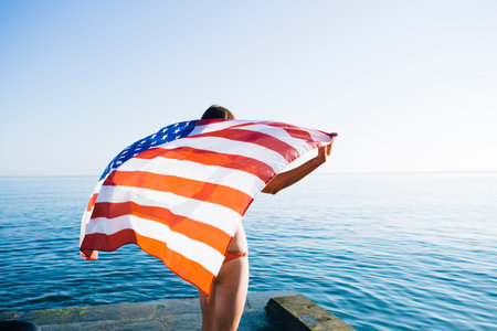 Rear view of unrecognizable young woman holding american waving flag at coastline against of sunny...