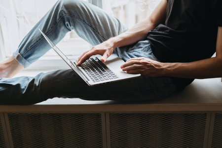 Cropped shot of a young male using his laptop while half-sitting on a windowsill.の写真素材