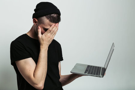 Close up studio shot of young guy in black hat and T-shirt holding laptop and showing facepalm...