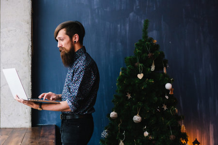Side view of a man standing near an xmas tree using a laptop