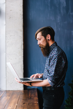 Side view of a standing bearded hipster using a laptopの写真素材
