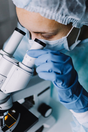 From above view of young scientist in work uniform looking at microscope.