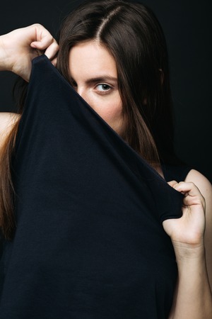 Young woman looking at camera and covering her face with her black cloth. Vertical studio shotの写真素材