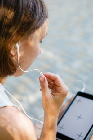 Back view of woman sitting on bench with headphones and using tablet outside.の写真素材
