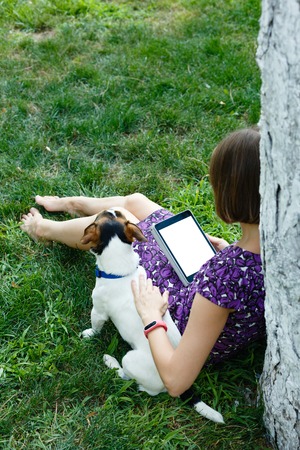 Anonymous woman in purple dress relaxing on green grass with tablet and her dog. Rear view. Crop shot with vertical orientation screen template.の写真素材