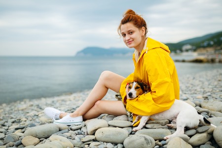 Side view of young woman in yellow raincoat posing on pebble beach with Jack Russell Terrier.の写真素材