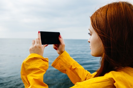 Young redhead woman in yellow raincoat posing on coastline of ocean taking photo of nature with smartphone.の写真素材
