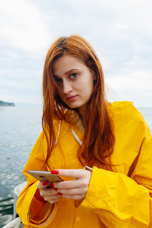 Portrait of young girl with red hair wearing bright coat and posing on ocean looking at camera.の写真素材