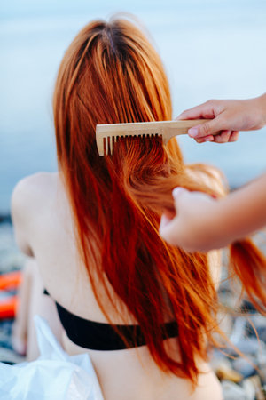 Back view of redhead woman in swimsuit sitting on beach and being brushed by anonymous person.の写真素材