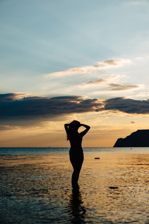 Black silhouette of woman posing in shallow water of ocean in tropics in sunset time.の写真素材