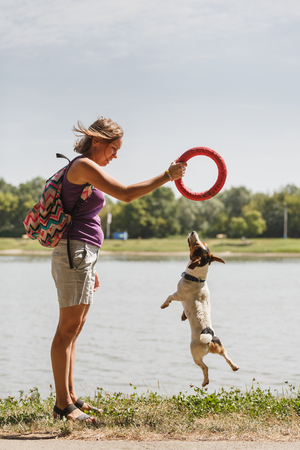 Side view of woman with backpack standing on background of lake in park and playing with dog.の写真素材